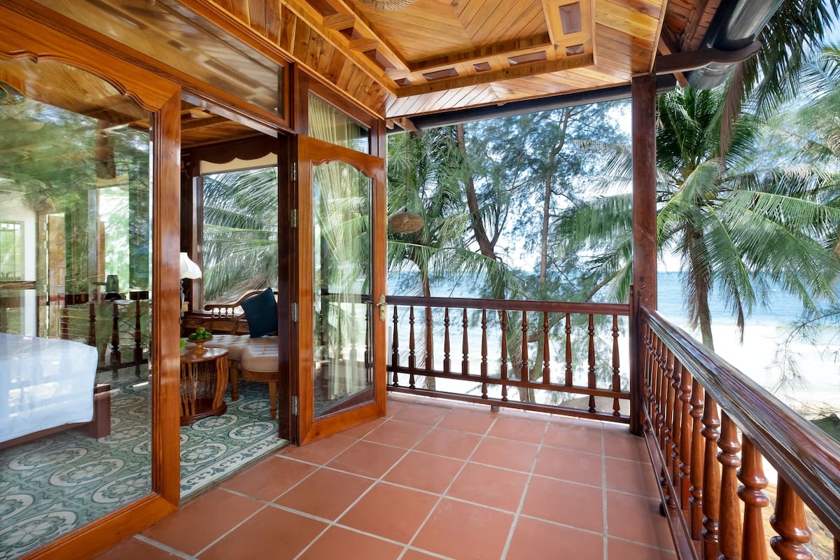 A wooden balcony with a tiled floor, a wooden railing, and a view of palm trees and the sea.