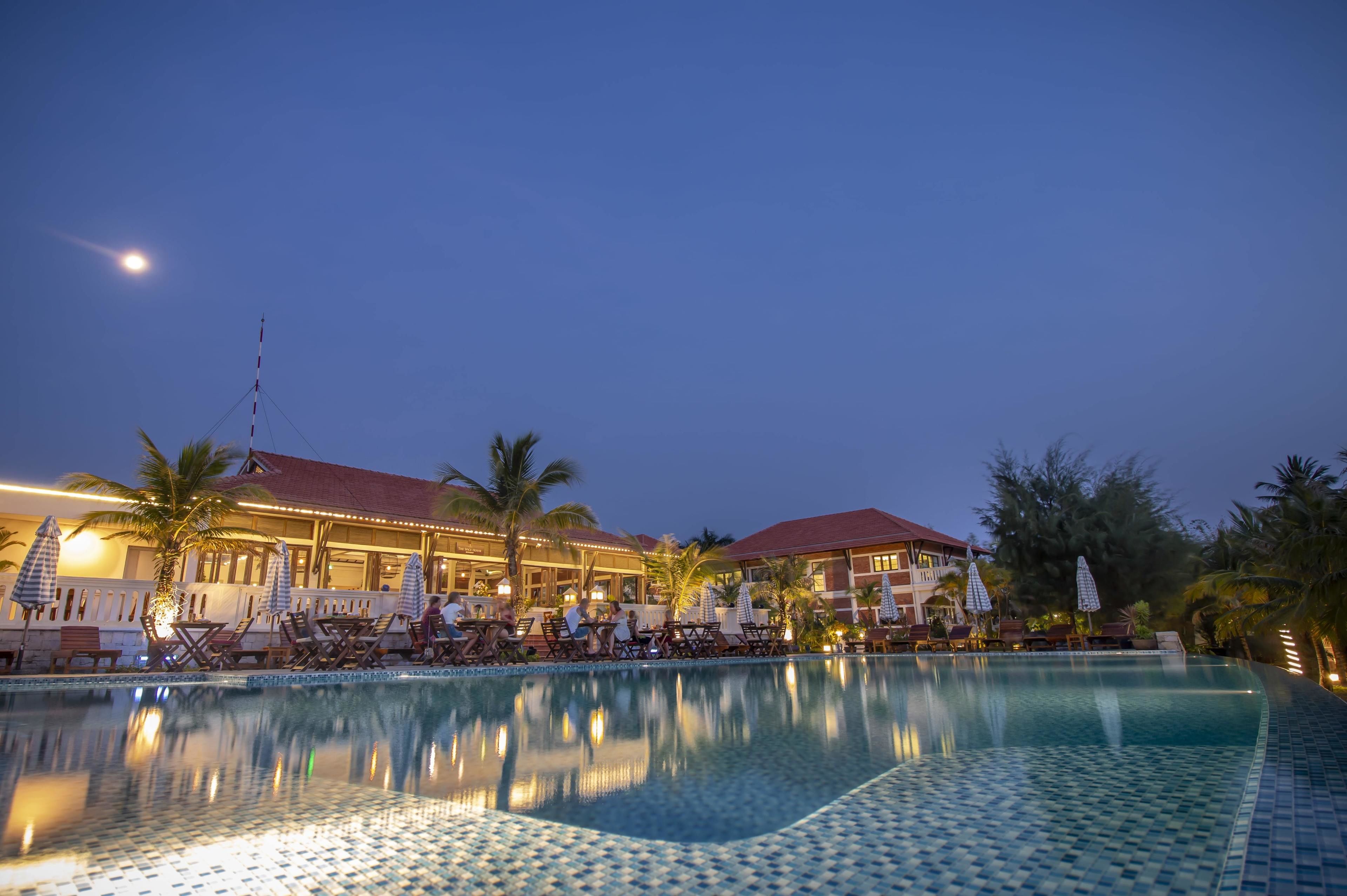 A hotel with a swimming pool, lounge chairs, umbrellas, and palm trees.