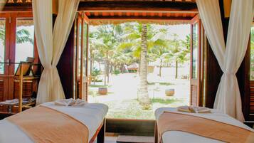 Two beds with crisp linens face a tranquil outdoor area with palm trees and a thatched-roof structure.