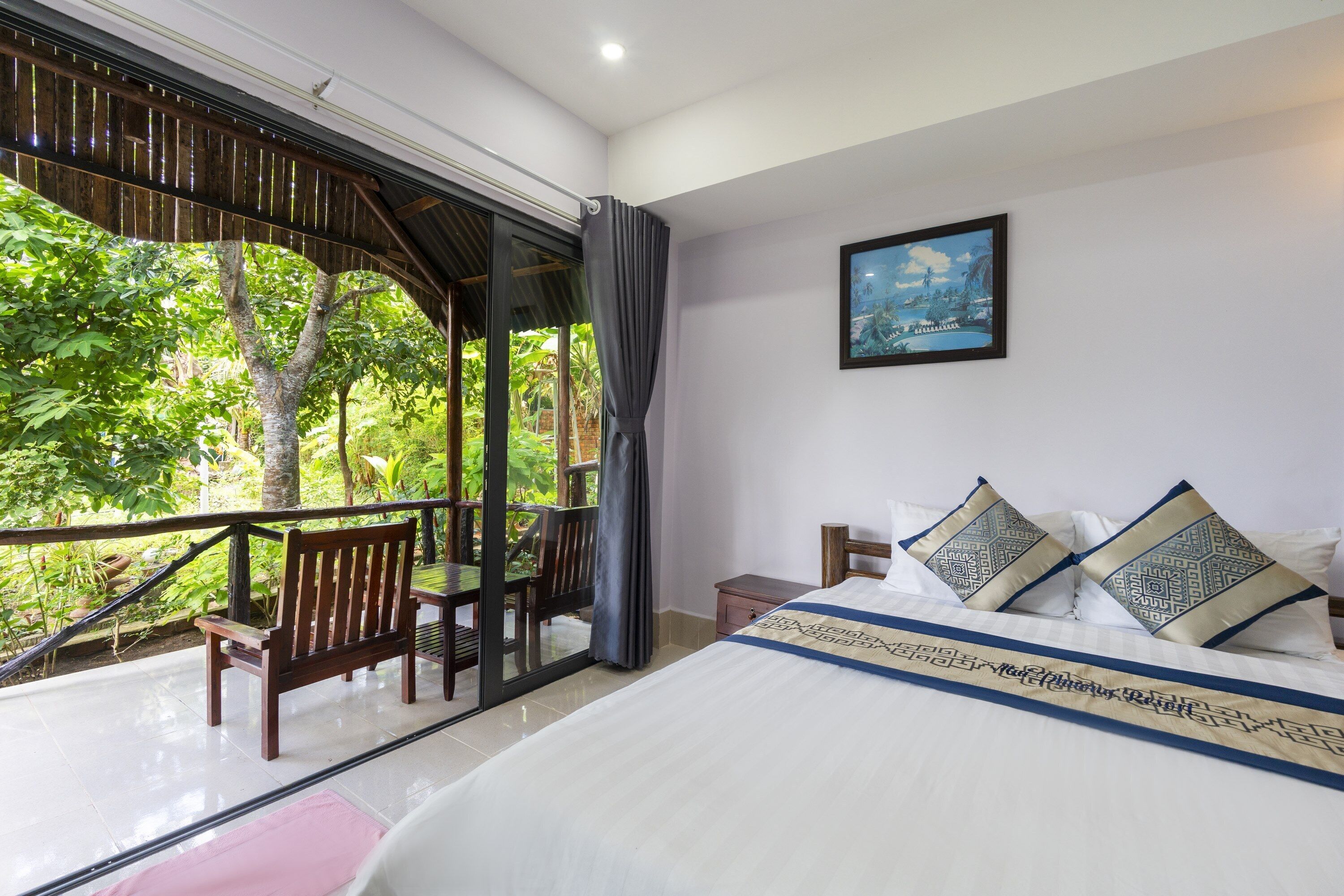 A bedroom with a large bed, a view of greenery through a glass door, a wooden chair, and a small table.
