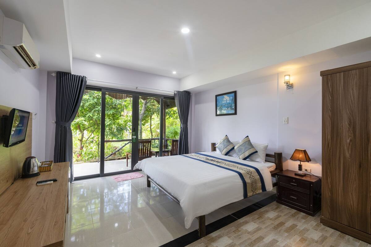 A bedroom with a large bed, a wooden nightstand, a wardrobe, and a sliding glass door leading to a balcony.