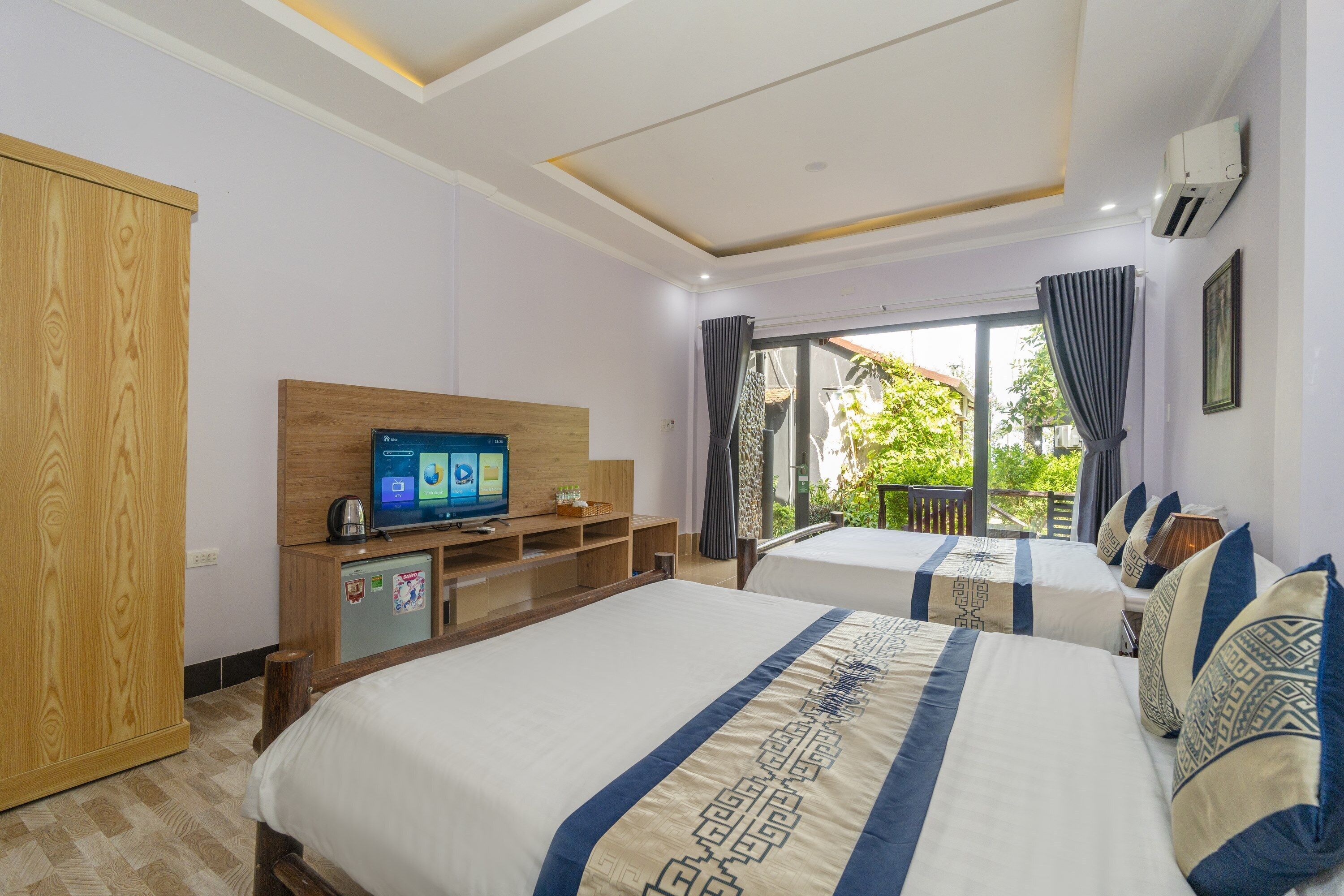 A hotel room with two beds, a TV, a minibar, and a balcony with a view of greenery.