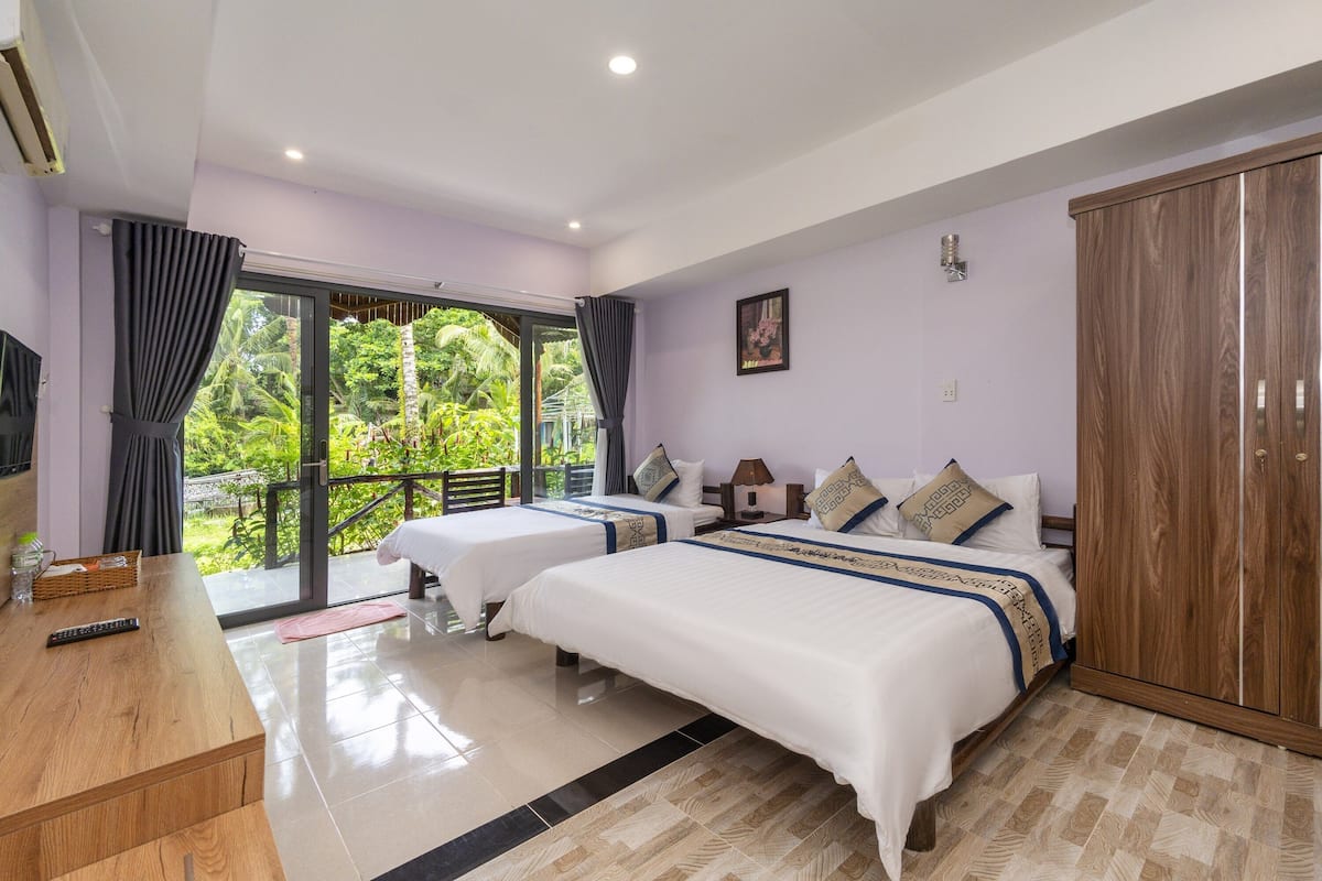 A hotel room with two beds, a wooden desk, a TV, and a balcony with a view of greenery.
