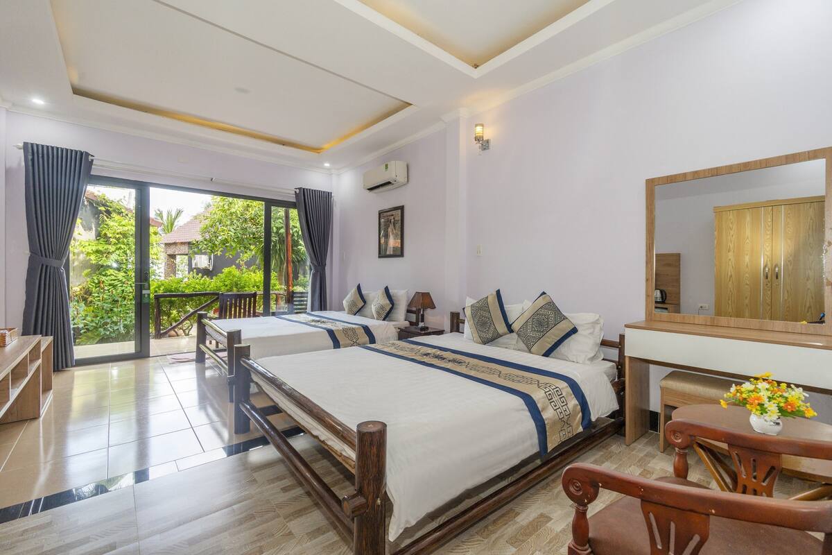 A hotel room with two beds, a wooden desk, a mirror, and a balcony with a view of greenery.