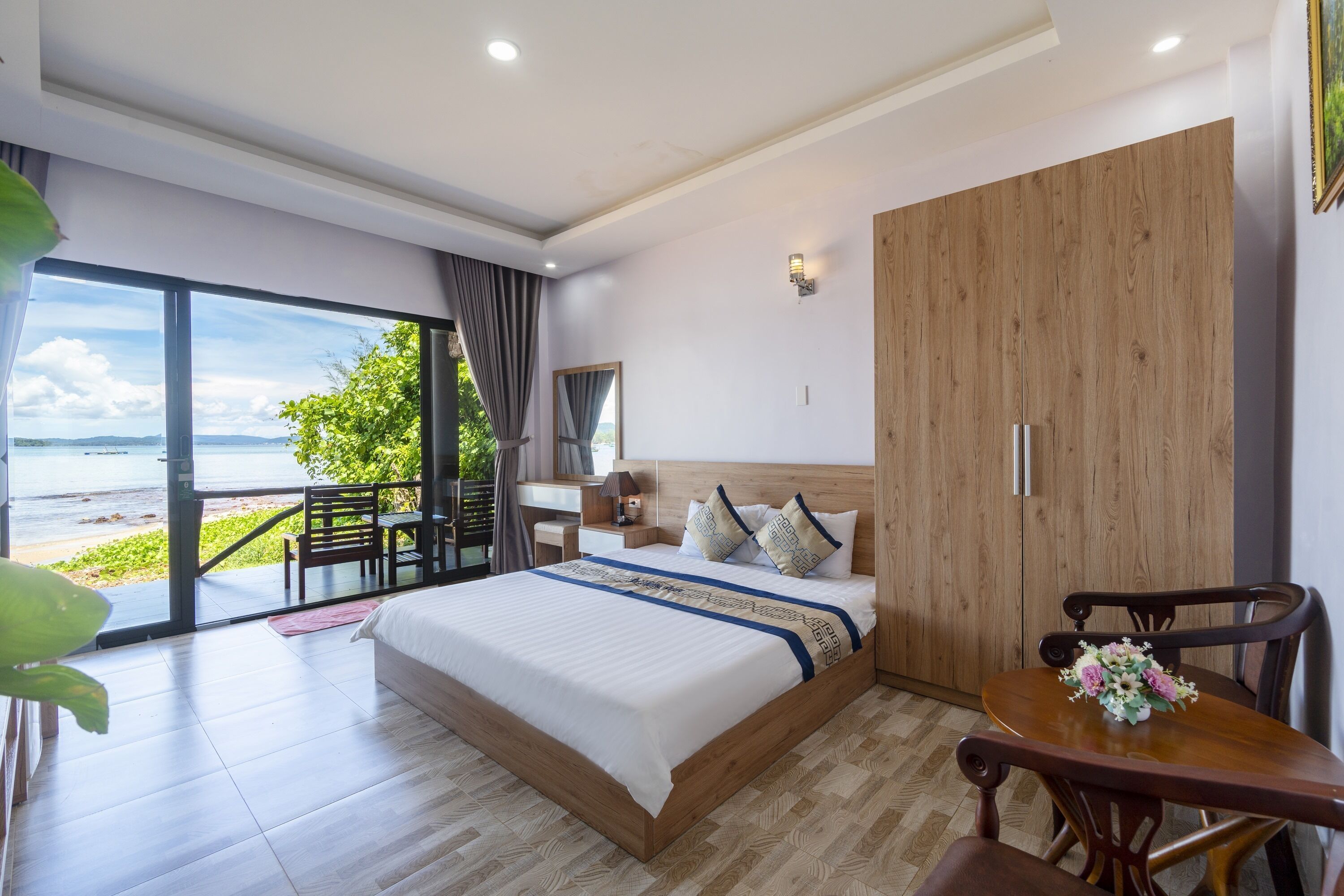 A bedroom with a large bed, a wooden wardrobe, a desk, and a chair. There is a view of the beach through a sliding door.