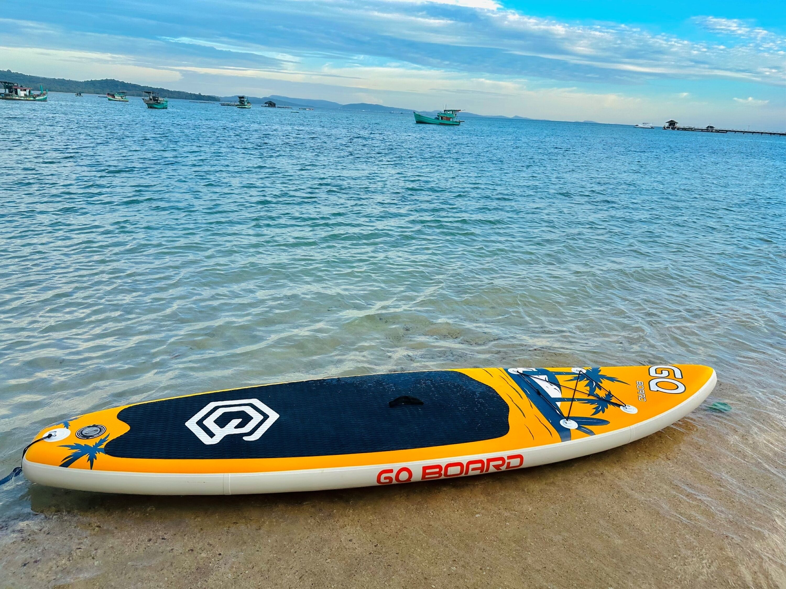 Una tavola Go Board da paddleboard su una spiaggia sabbiosa con acqua calma e barche sullo sfondo.