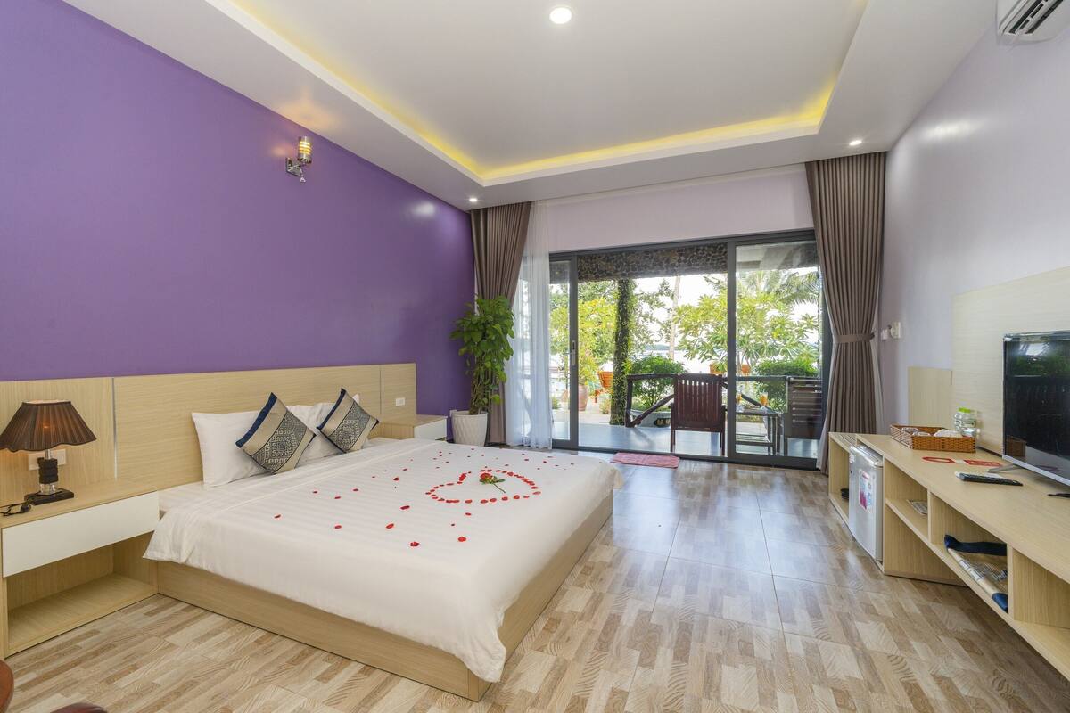 A modern bedroom with a large bed, a TV, and a balcony with a view of greenery.
