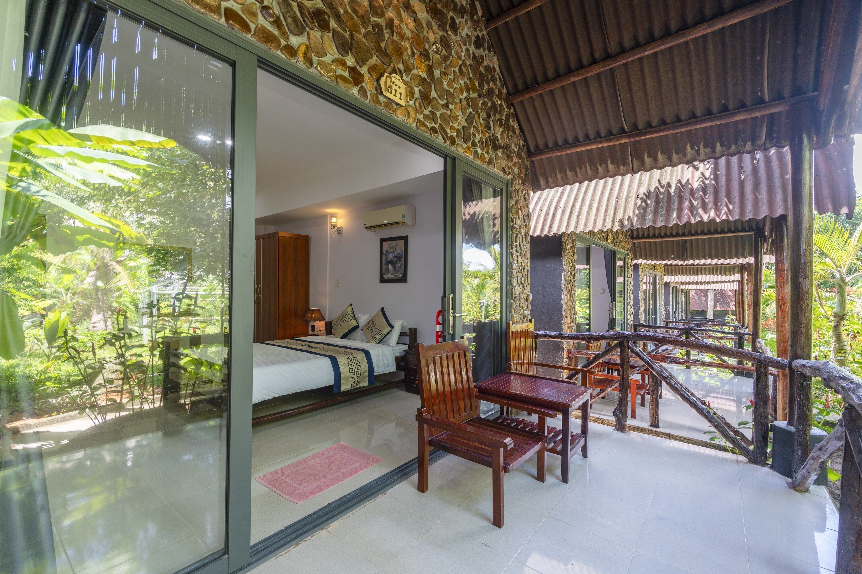 A room with a bed, a wooden chair, and a table. There is a glass door leading to an outdoor area with plants.
