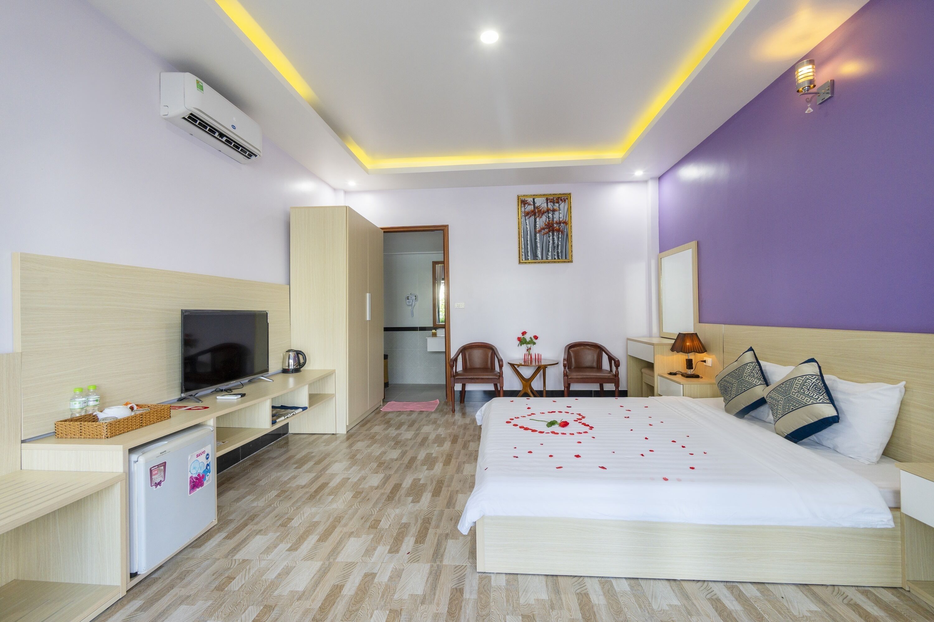 A hotel room with a bed, a TV, a desk, a mini-fridge, a chair, and a small table with a lamp.