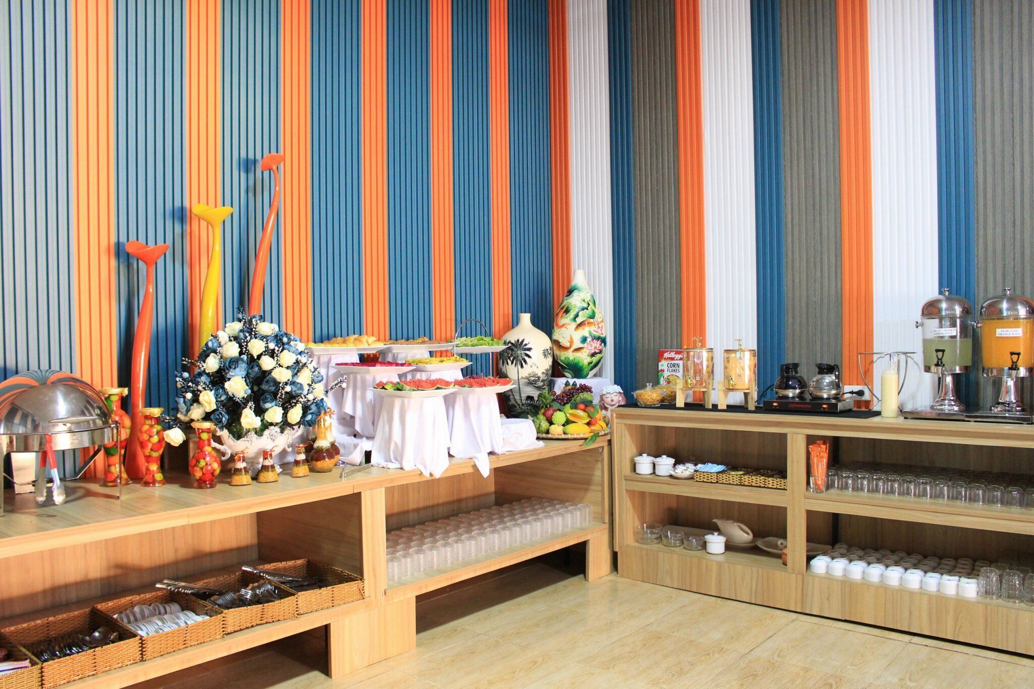 Een buffetopstelling met diverse gerechten en dranken, een decoratieve bloemstuk en een gestreepte wand op de achtergrond.