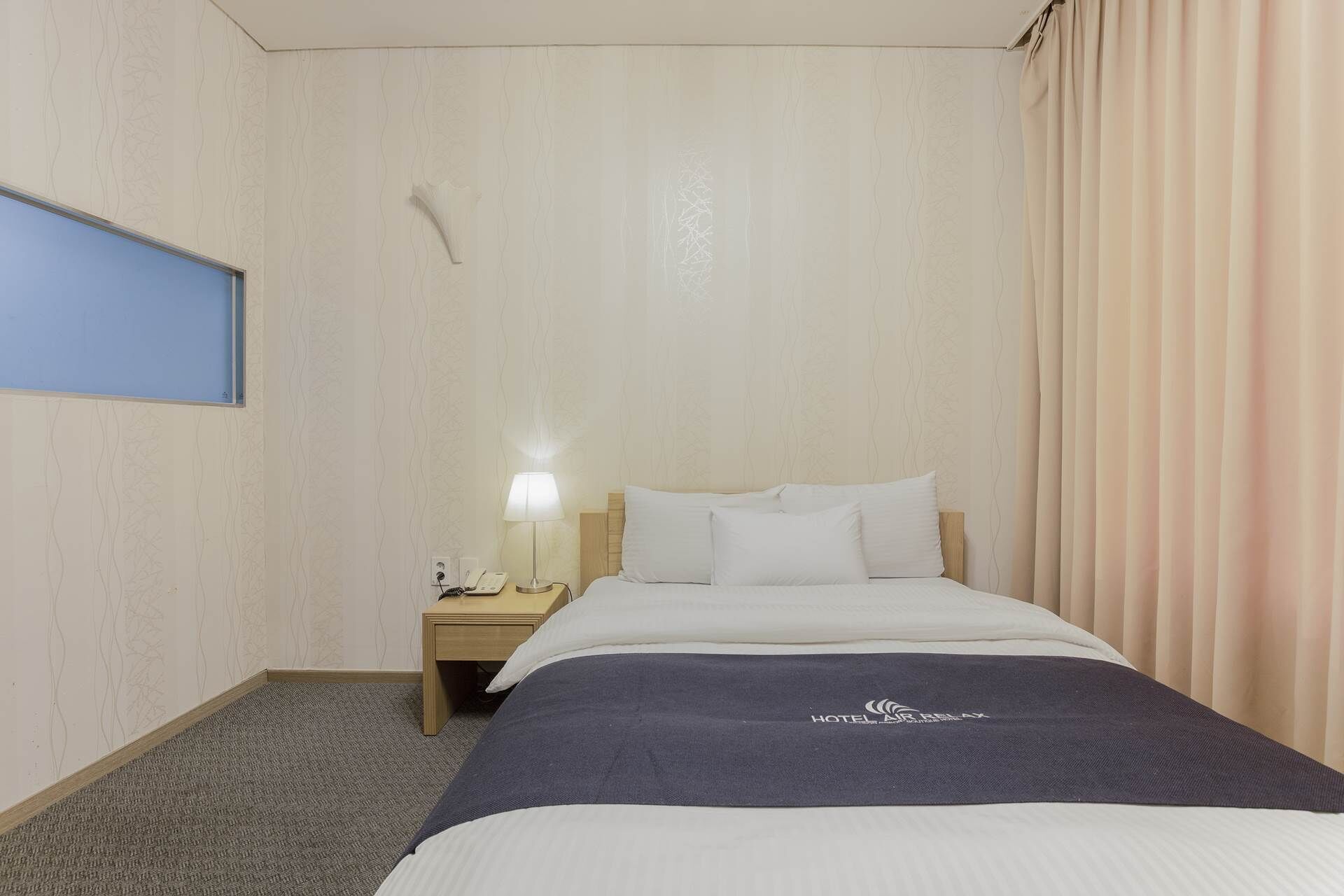 Una habitación de hotel con una cama, una mesita de noche con lámpara, un teléfono en la pared y una ventana con cortina.