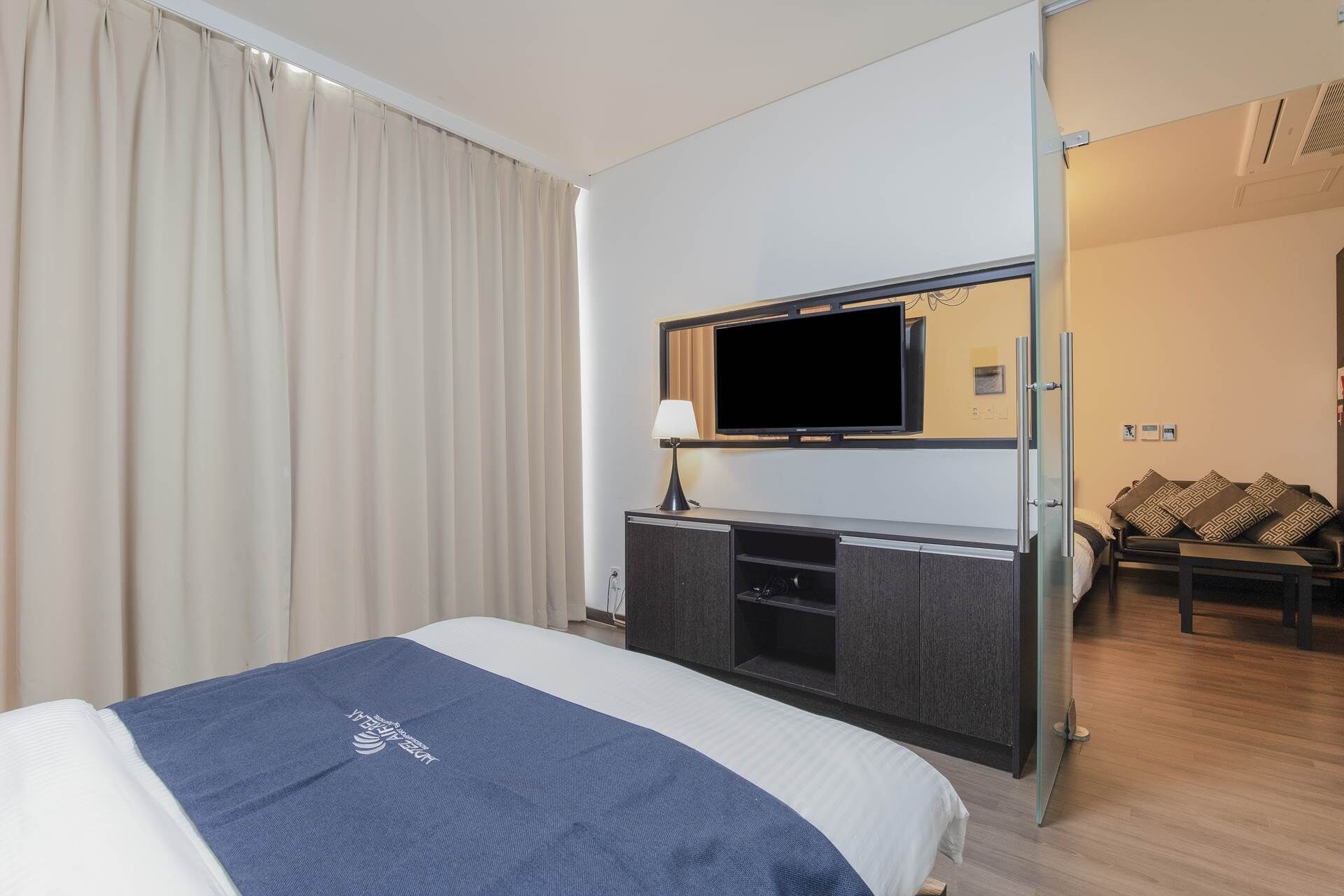 A hotel room with a bed, a desk with a flat-screen TV, a lamp, and a seating area with cushions.