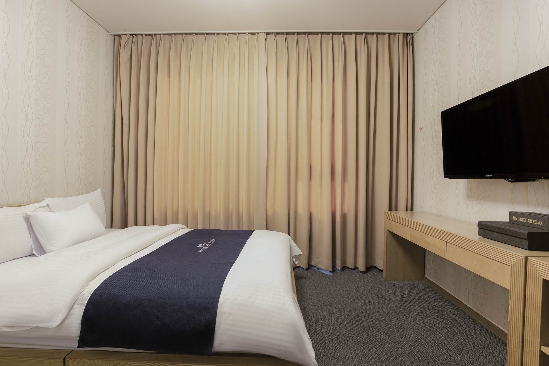 Ein Hotelzimmer mit Bett, Flachbildfernseher, Schreibtisch und großem Fenster mit Vorhängen.