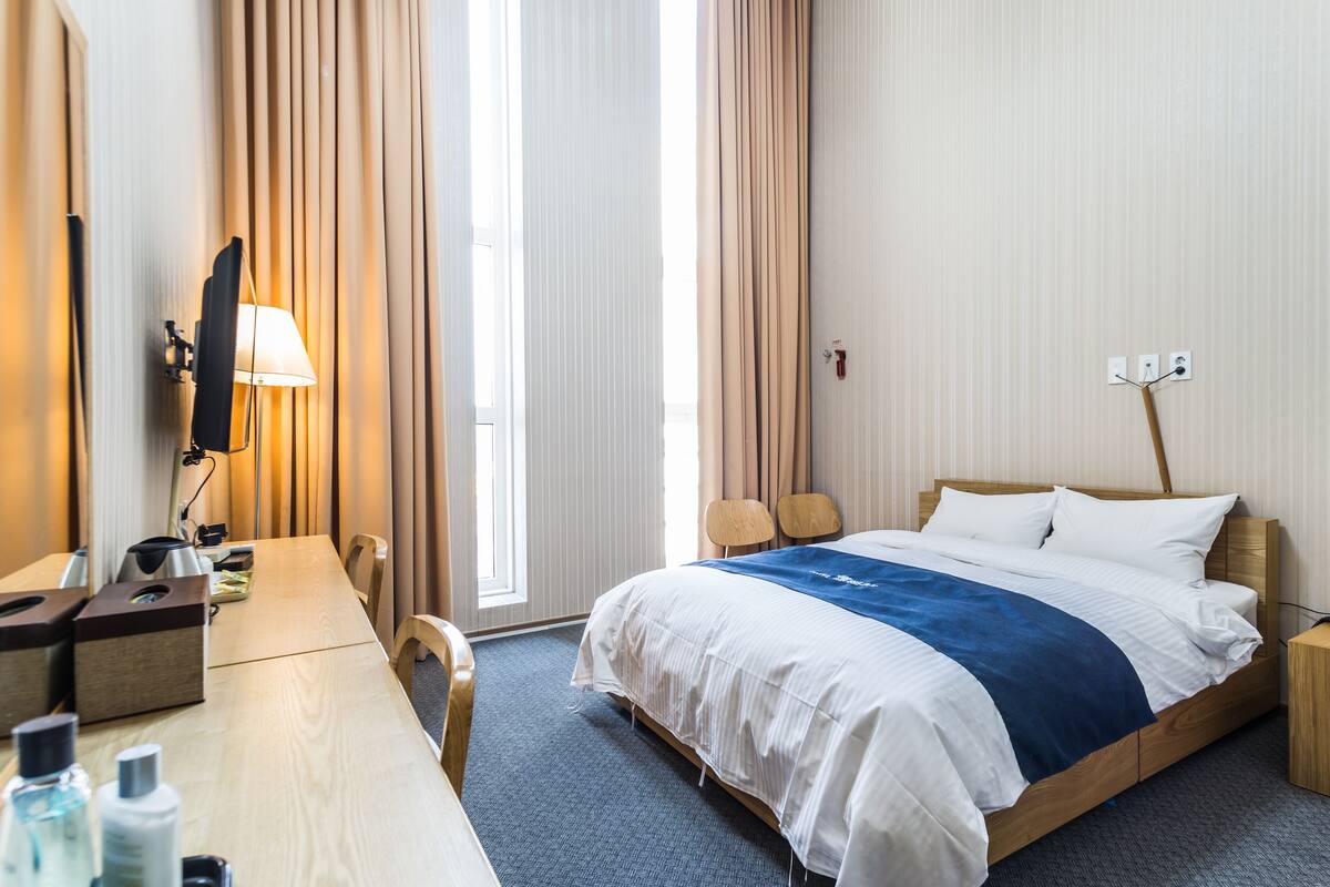 A hotel room with a bed, bedside table, lamp, and a view of the outdoors.
