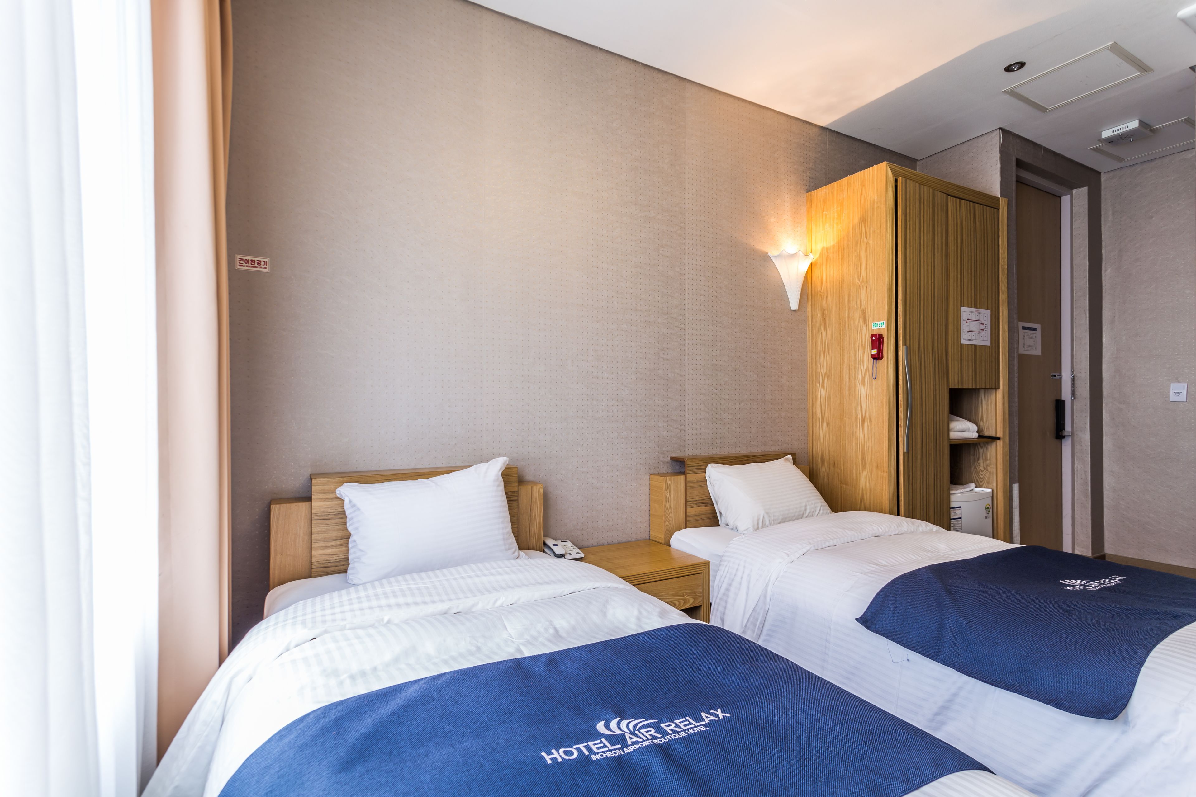 Habitación de hotel con dos camas, un armario de madera, una mesita de noche con teléfono, y una lámpara fijada a la pared.