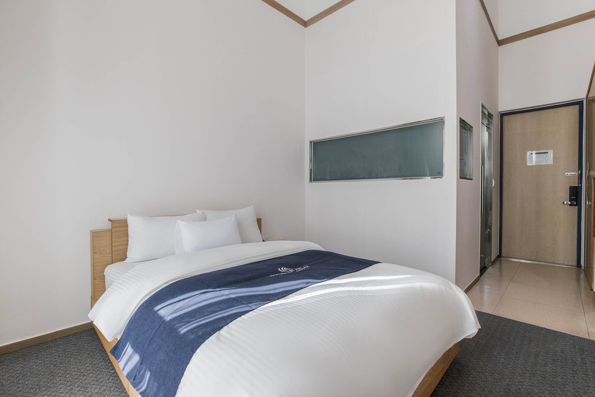A hotel room with a bed, a wooden headboard, a blue and white blanket, white pillows, a small window, and a door.