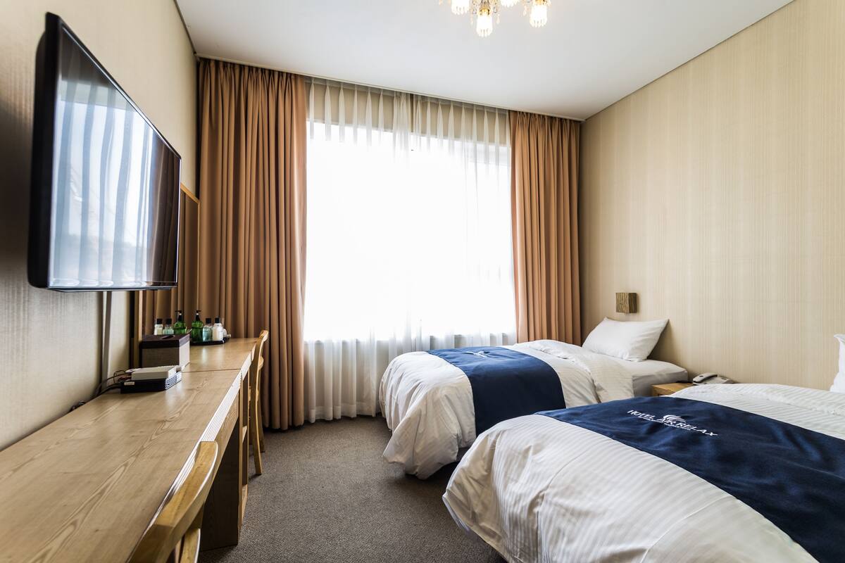 A hotel room with two beds, a desk, a television, and a large window with curtains.