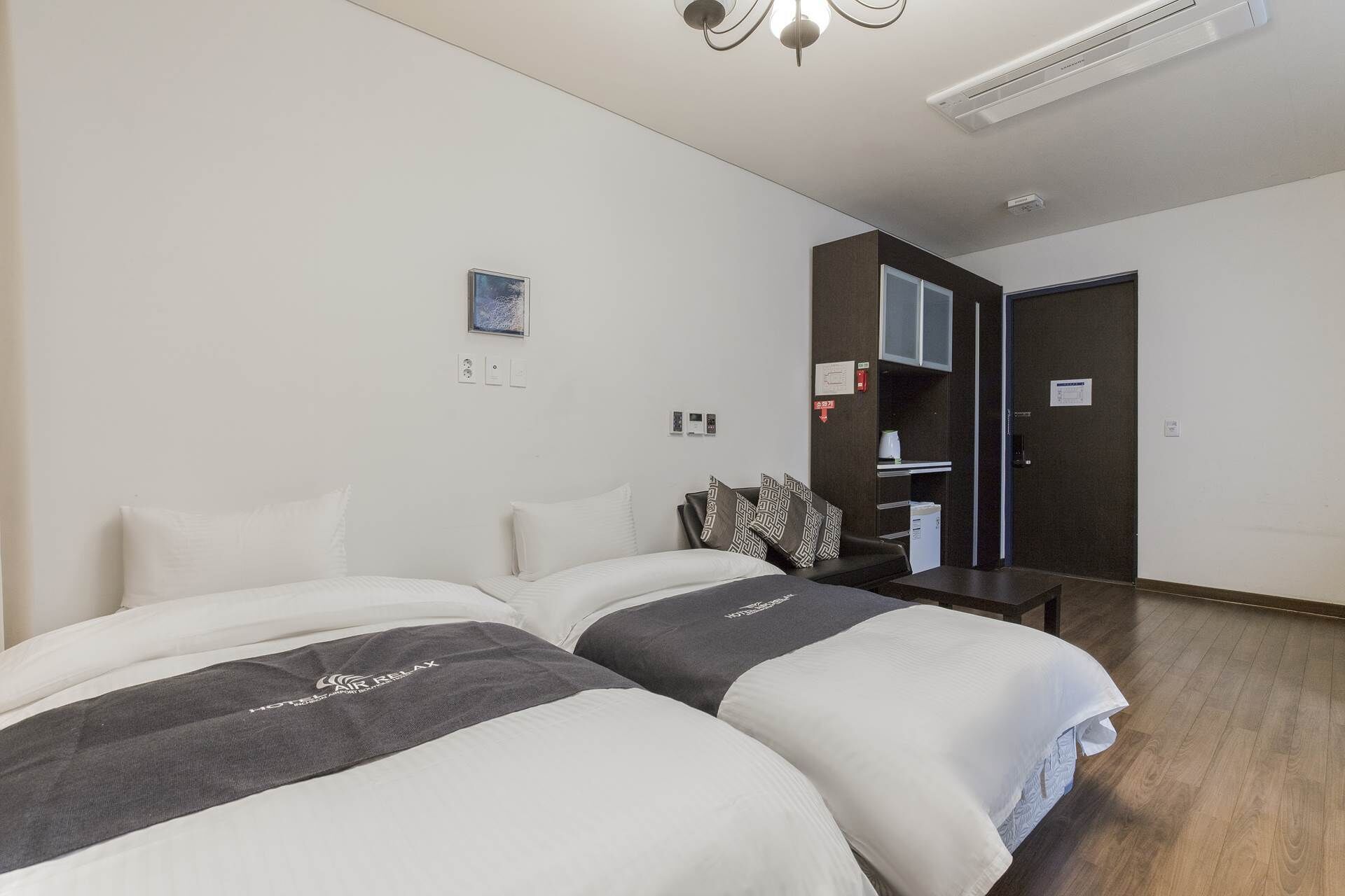 Habitación de hotel con dos camas, un escritorio, una silla, una mesita, una estantería y una puerta.