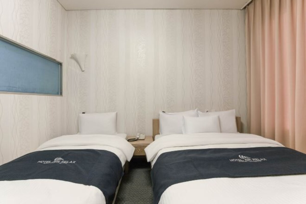 Habitación de hotel con dos camas, cada una con cubrecama azul oscuro y almohadas blancas. Entre las camas hay una mesita de noche de madera con un teléfono y una lámpara pequeña. La habitación tiene una pared de madera de color claro y una ventana con marco azul.
