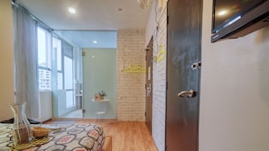A hotel room with a wooden floor, a bed with a patterned bedspread, a small table with a vase and a basket, a brick wall, and a door with a towel rack. - Snooze Too (Brinchang)