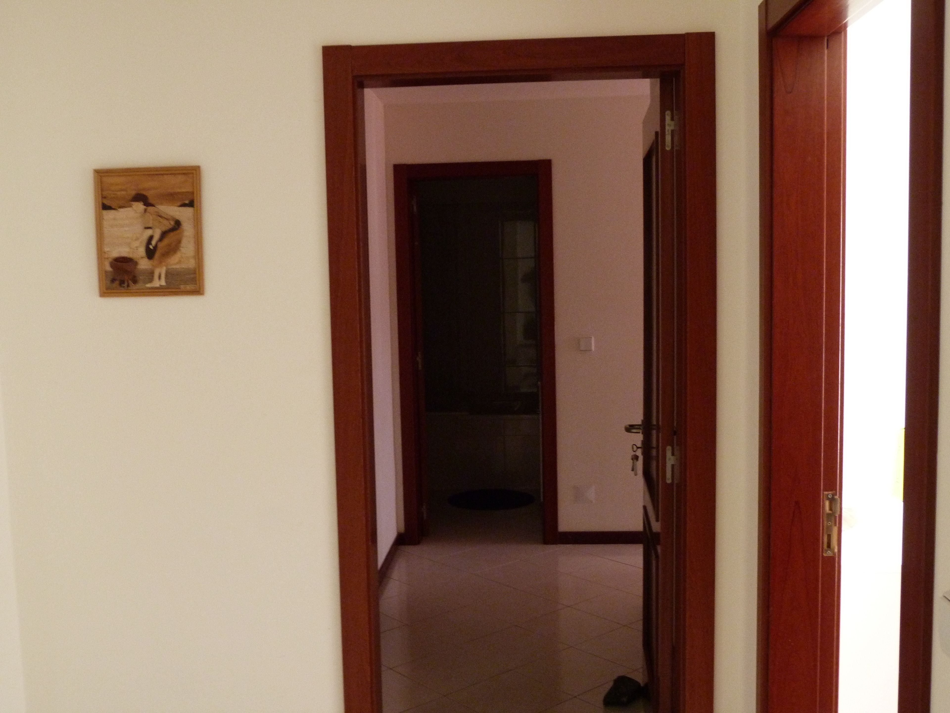 A hallway with a wooden door leading to a dark room. A small framed picture hangs on the wall to the left.