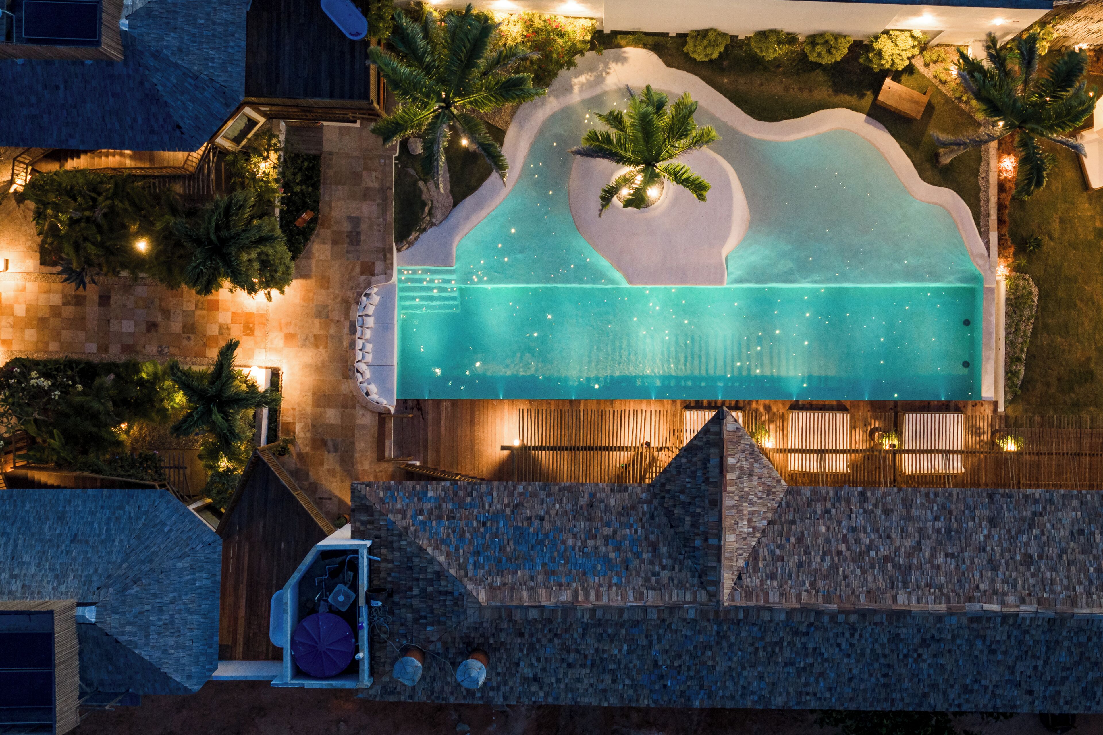 Vue aérienne d’un complexe hôtelier avec une piscine, des palmiers et une terrasse en bois.