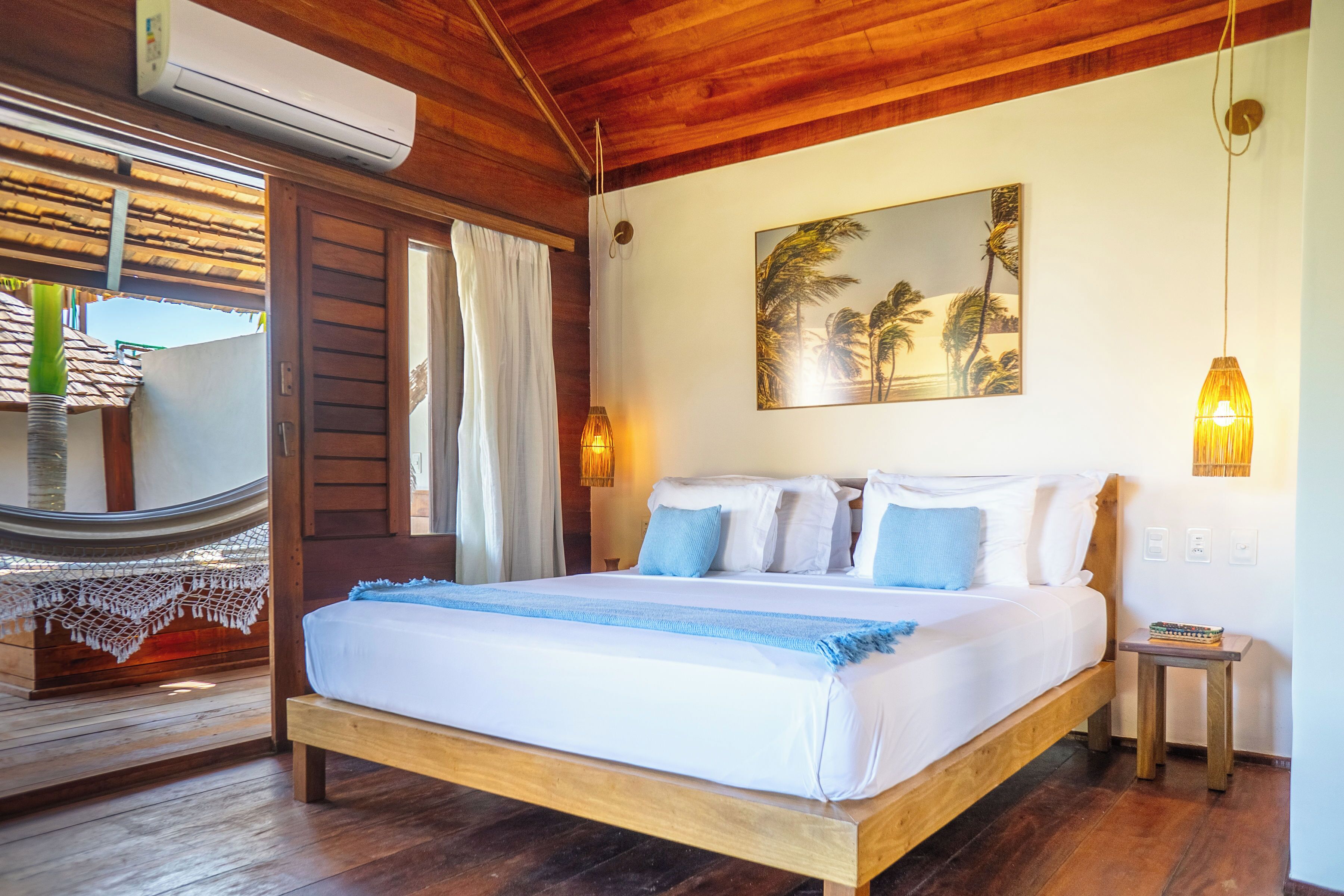 Een bed met houten frame, witte beddengoed en blauwe accenten, een hangmat buiten en een wandairconditioning.