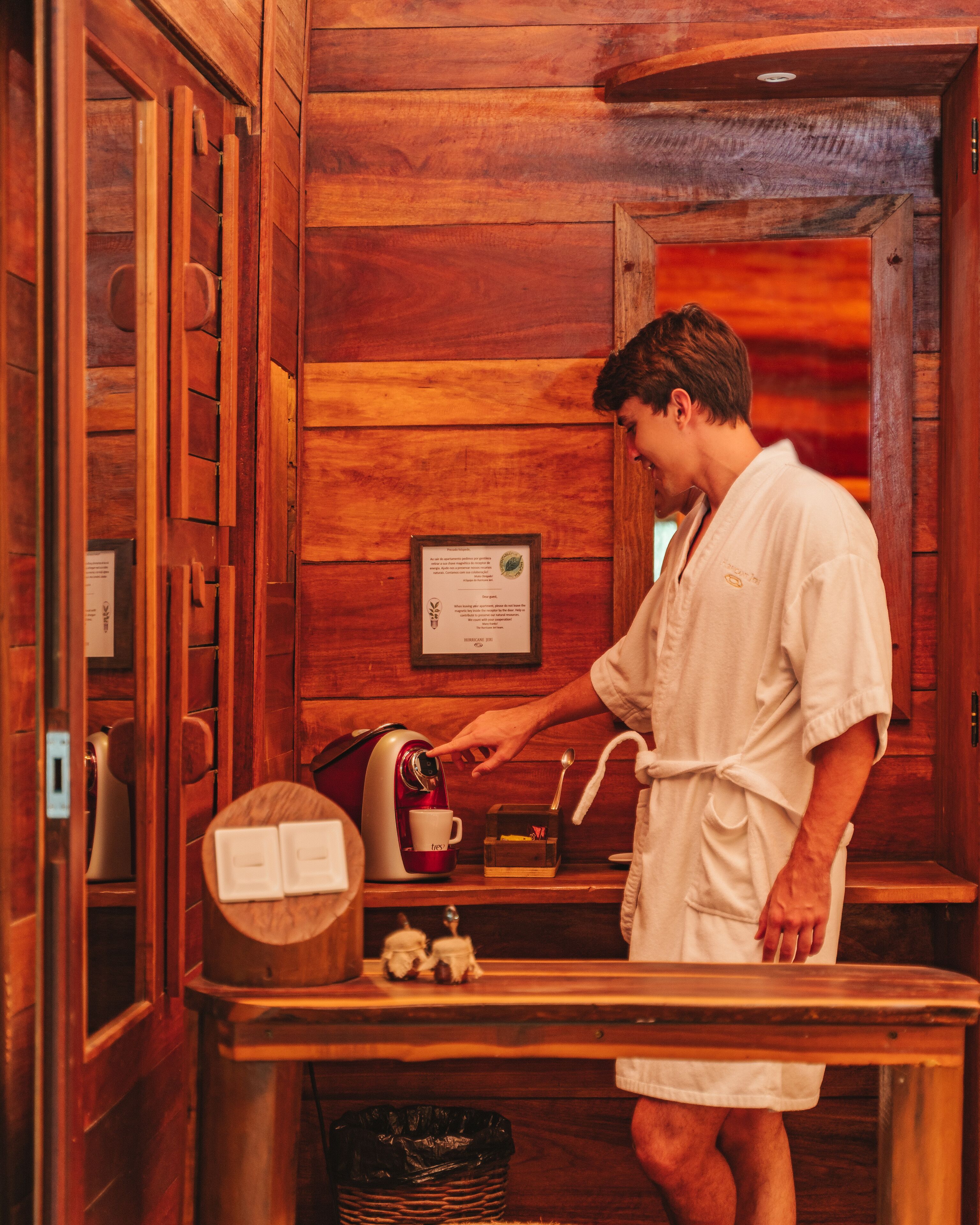 Une personne vêtue d’une robe blanche prépare du café dans une pièce aménagée comme un chalet en bois, avec une table en bois, une machine à café et un certificat encadré accroché au mur.