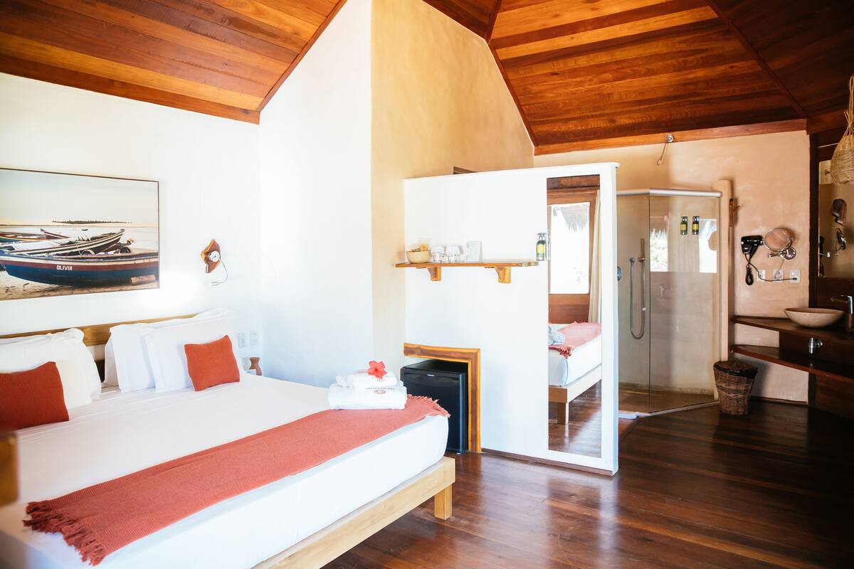 A bedroom with a bed, a wooden ceiling, a painting, a small table with a lamp, and a bathroom visible through an open door.