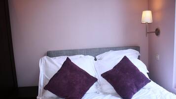 Un lit bien fait, avec du linge de lit blanc et deux coussins violets, une tête de lit grise et un luminaire fixé au mur.