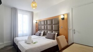 A hotel room with a large bed, a headboard with built-in lighting, a window with curtains, and a door with a handle. - Hotel de France (Paris)