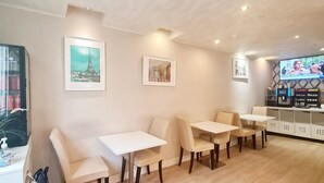 A restaurant with white tables and chairs, framed pictures on the walls, and a television screen displaying a program. - Hotel de France (Paris)