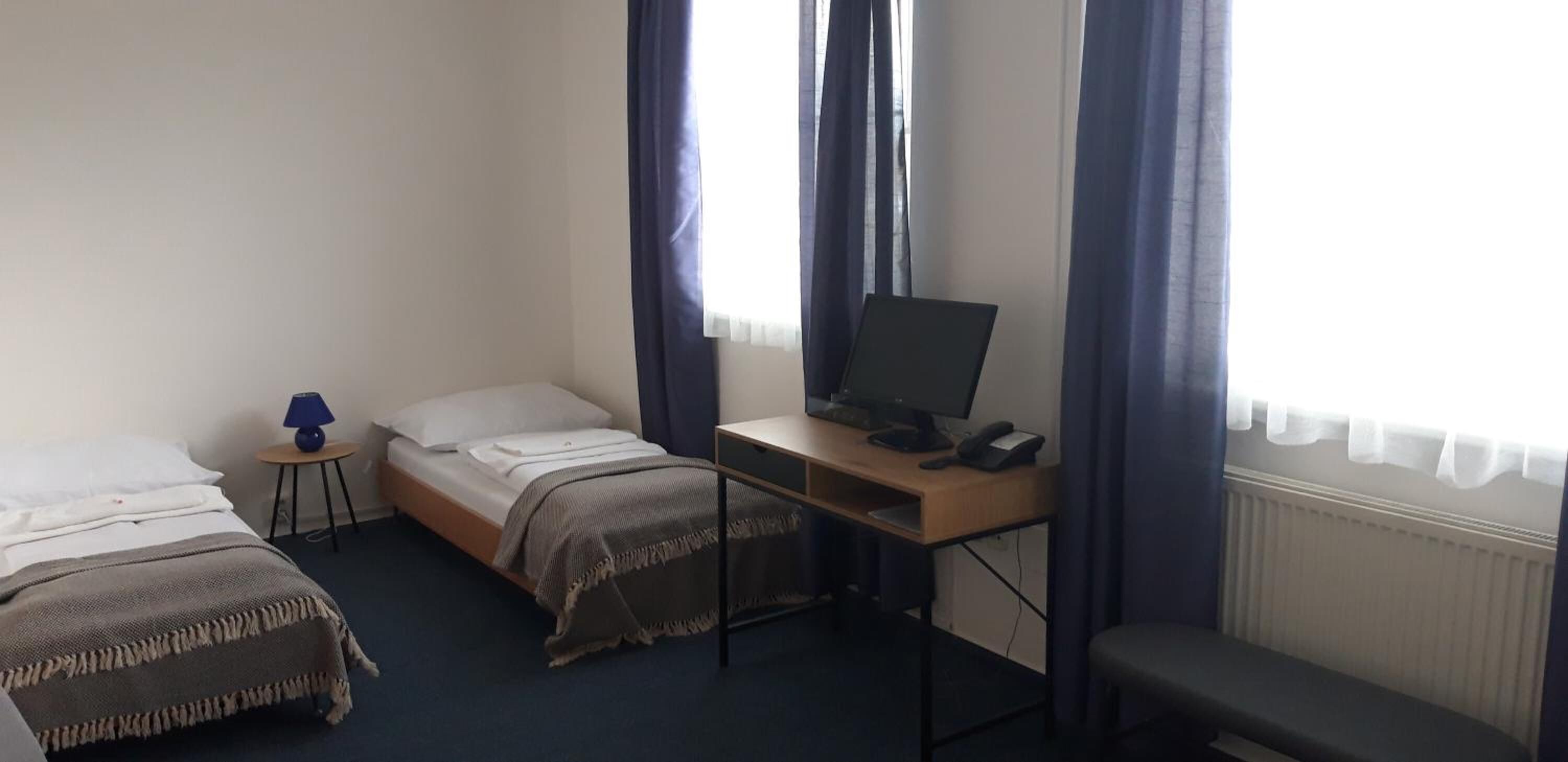 A small hotel room with two single beds, a desk with a computer, a lamp, and a window with curtains.