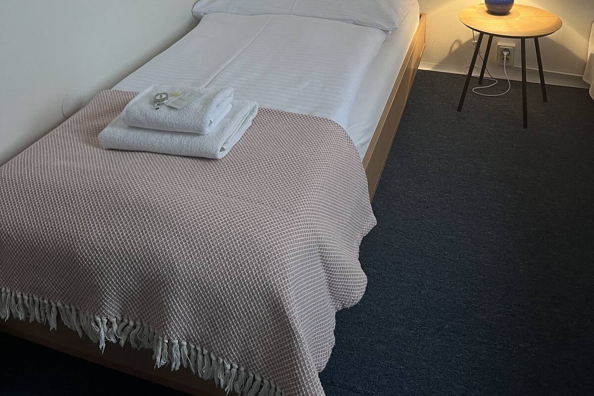 A single bed with a white mattress and a pink blanket, a small round table with a lamp, and a carpeted floor.