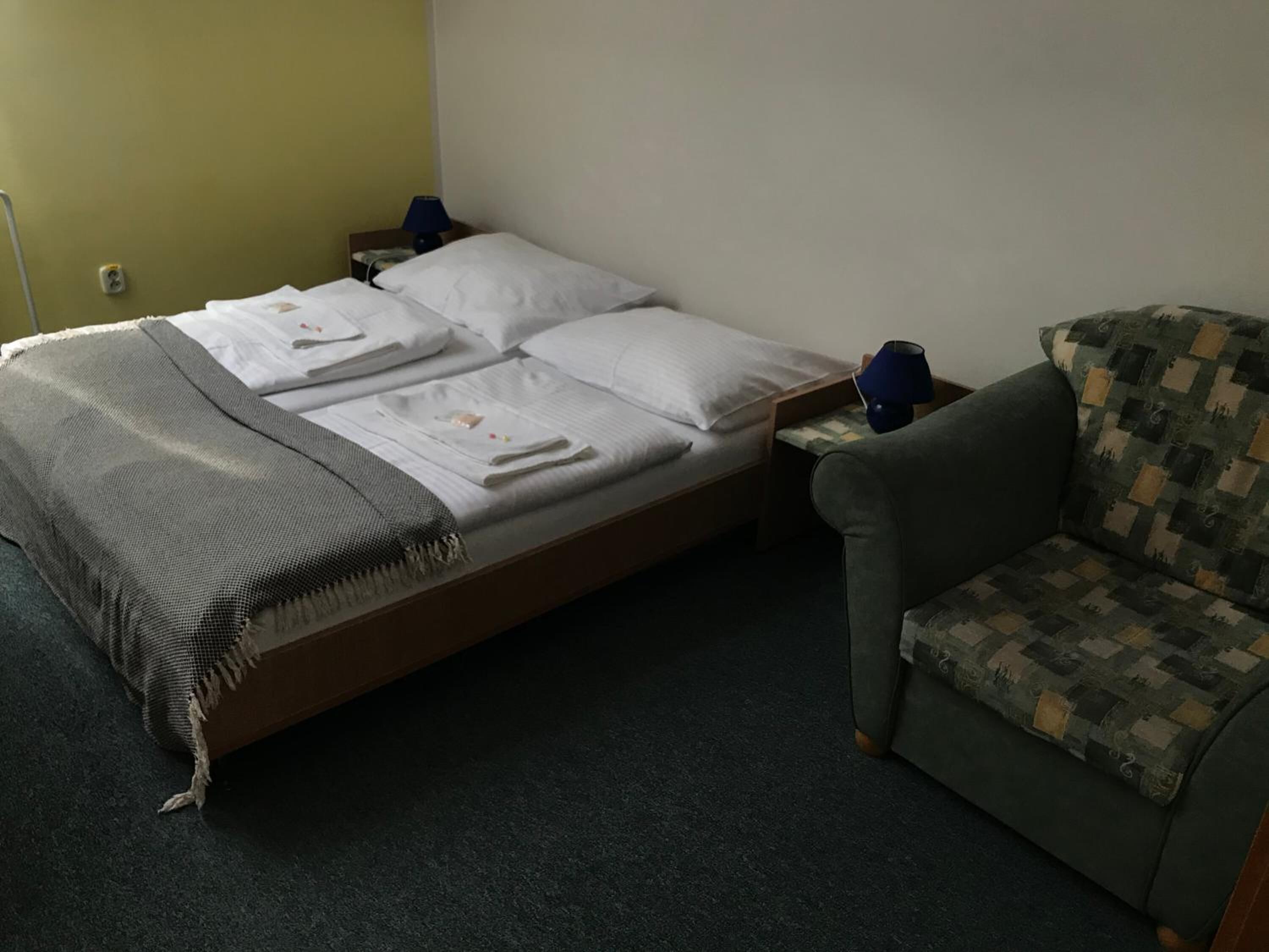 A single bed with white linens and a gray throw blanket, a small table with a blue lamp, and a patterned armchair.