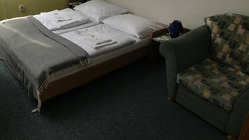 A single bed with white linens and a gray throw blanket, a small table with a blue lamp, and a patterned armchair.