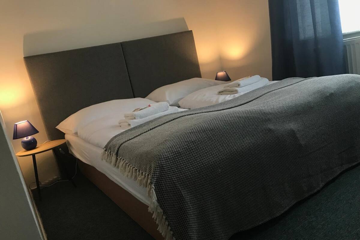 A neatly made bed with a grey and white striped blanket, two pillows, and a bedside lamp on a wooden nightstand.