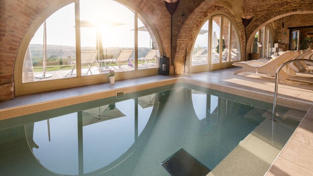 Una piscina interior con ventanales arqueados, sillas de descanso y vistas al paisaje.