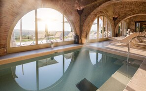 Una piscina interior con ventanales arqueados, sillas de descanso y vistas al paisaje.
