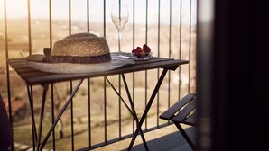 Un balcón con una mesa lista para dos personas, que incluye un sombrero de paja, una copa de vino y un tazón de fresas.