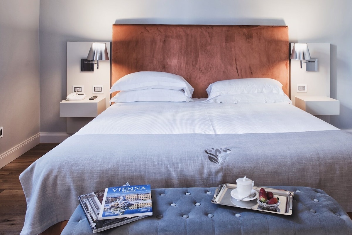Een netjes opgemaakt bed met een houten hoofdbord, twee bedlampen, een nachtkastje met een telefoon en afstandsbediening, en een dienblad met een kopje en aardbeien op een poef.