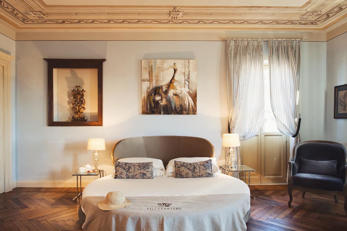 Een hotelkamer met een groot bed, twee nachtkastjes met lampen, een decoratief wandobject, een stoel en uitzicht buiten door gordijnen.