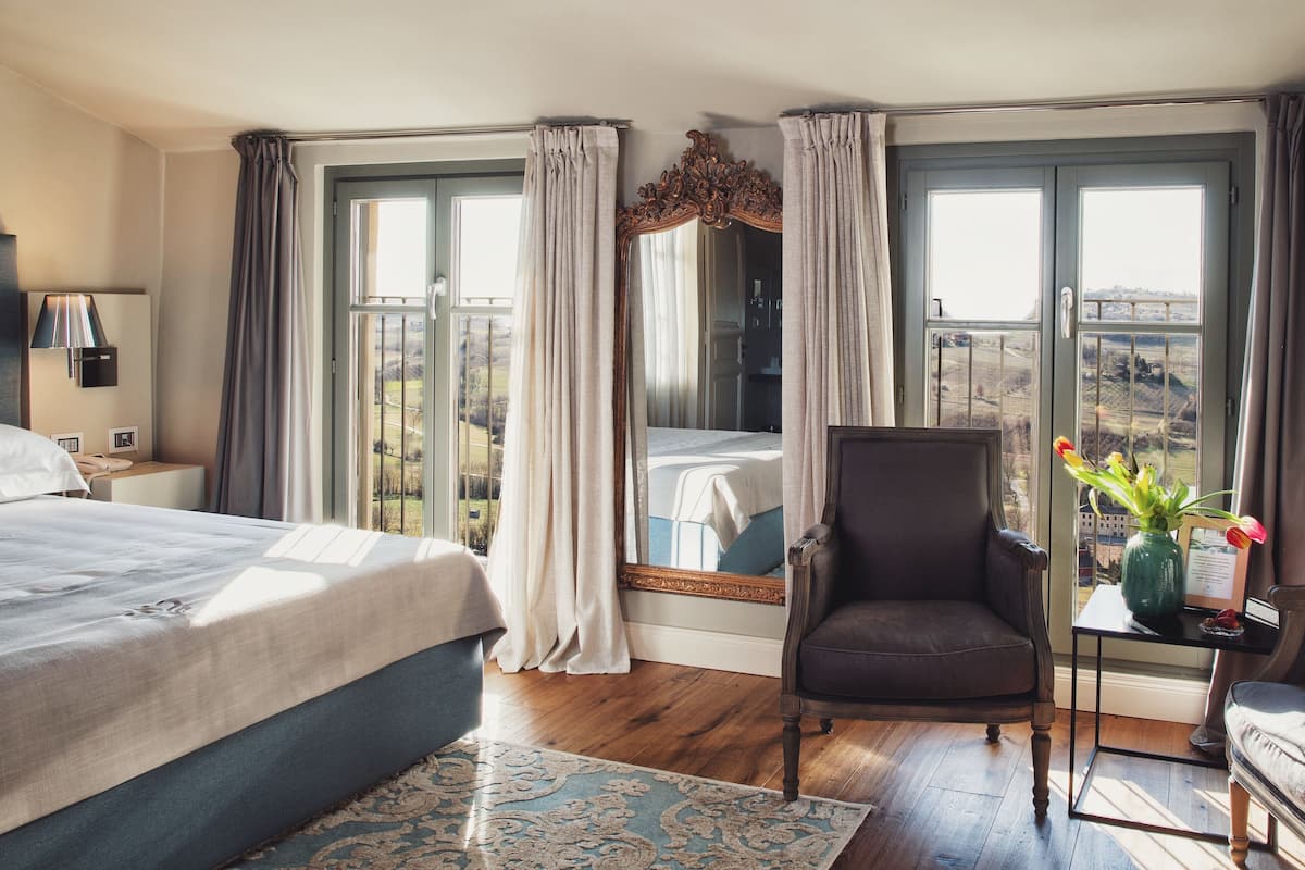 Een hotelkamer met een bed, een stoel, een klein tafeltje met een bloevas en uitzicht op het landschap door grote ramen.