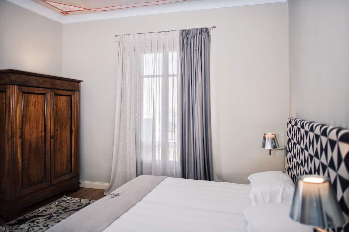 Een slaapkamer met een houten kledingkast, een bed met witte lakens, een gestreept hoofdeinde, een nachtkastje met een lamp en een raam met dunne gordijnen.