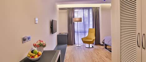 Quarto de hotel moderno com piso de madeira, uma mesa de jantar com uma fruteira, uma revista, uma poltrona amarela, uma TV e uma cama com vista através de cortinas finas.