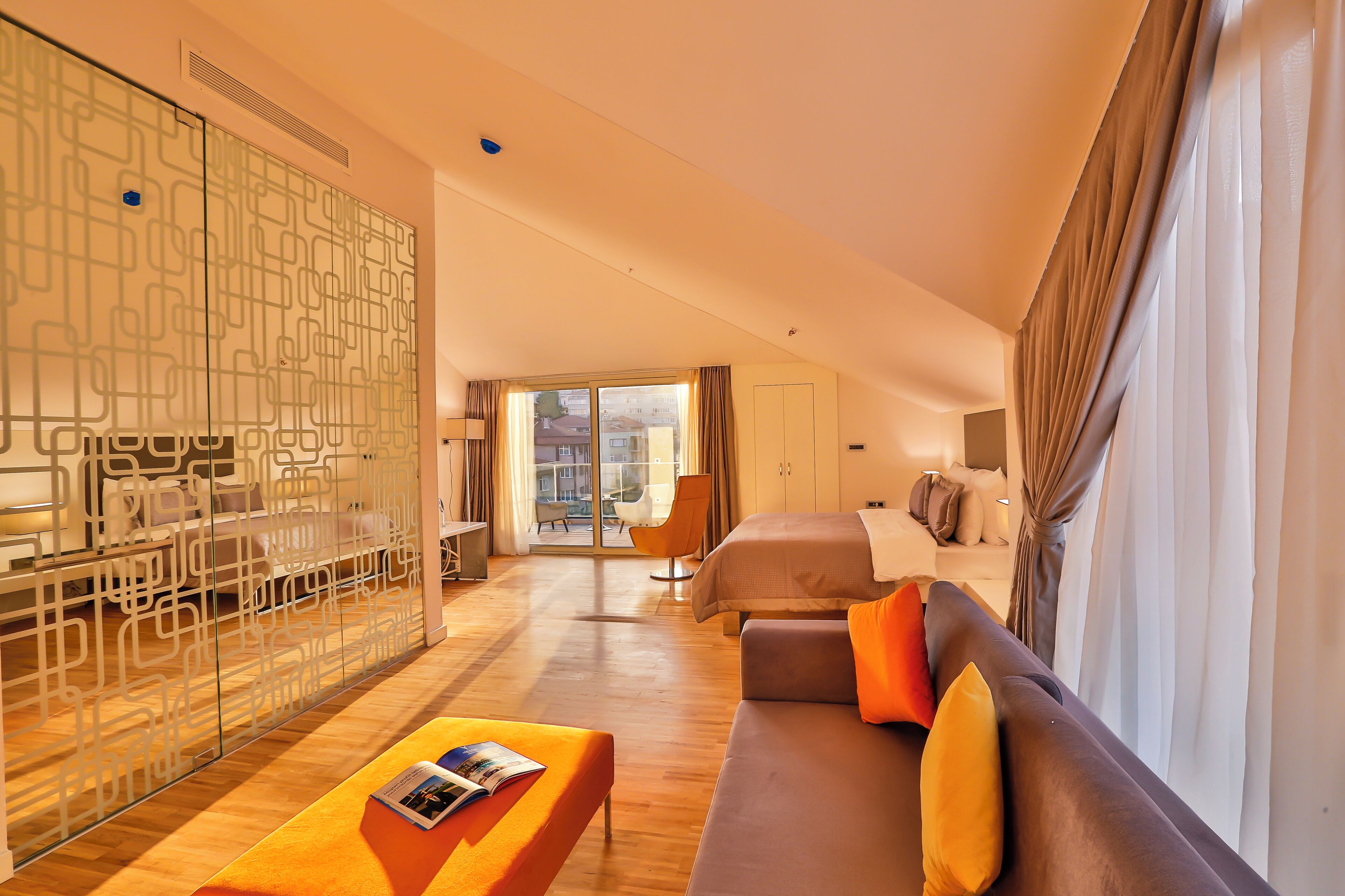 A modern hotel room with a wooden floor, a sofa with orange cushions, a bed, a glass partition, and a view of the city through large windows.