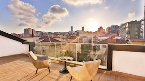 Een balkon met twee stoelen en een tafel, met uitzicht op een stadsbeeld met gebouwen en een heldere lucht.