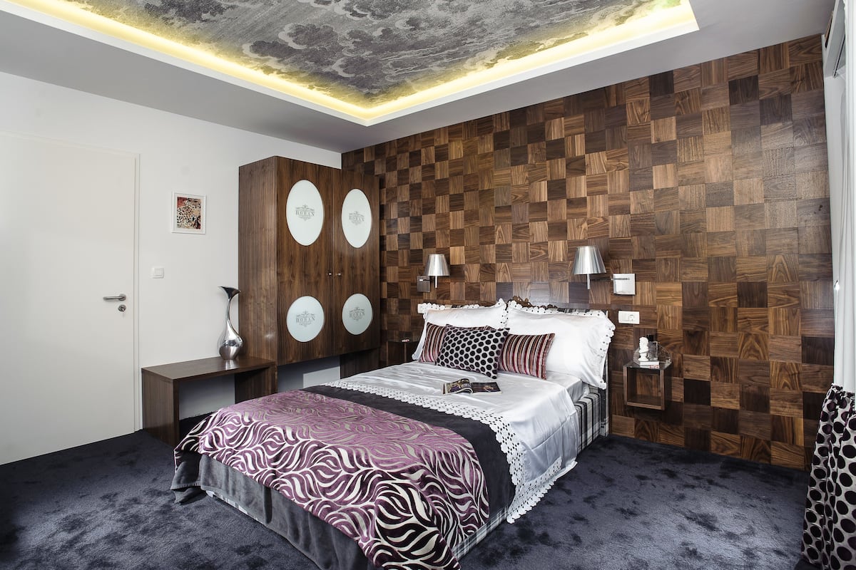 A modern bedroom with a wooden wall panel, a bed with patterned bedding, and a ceiling with a cloud design.