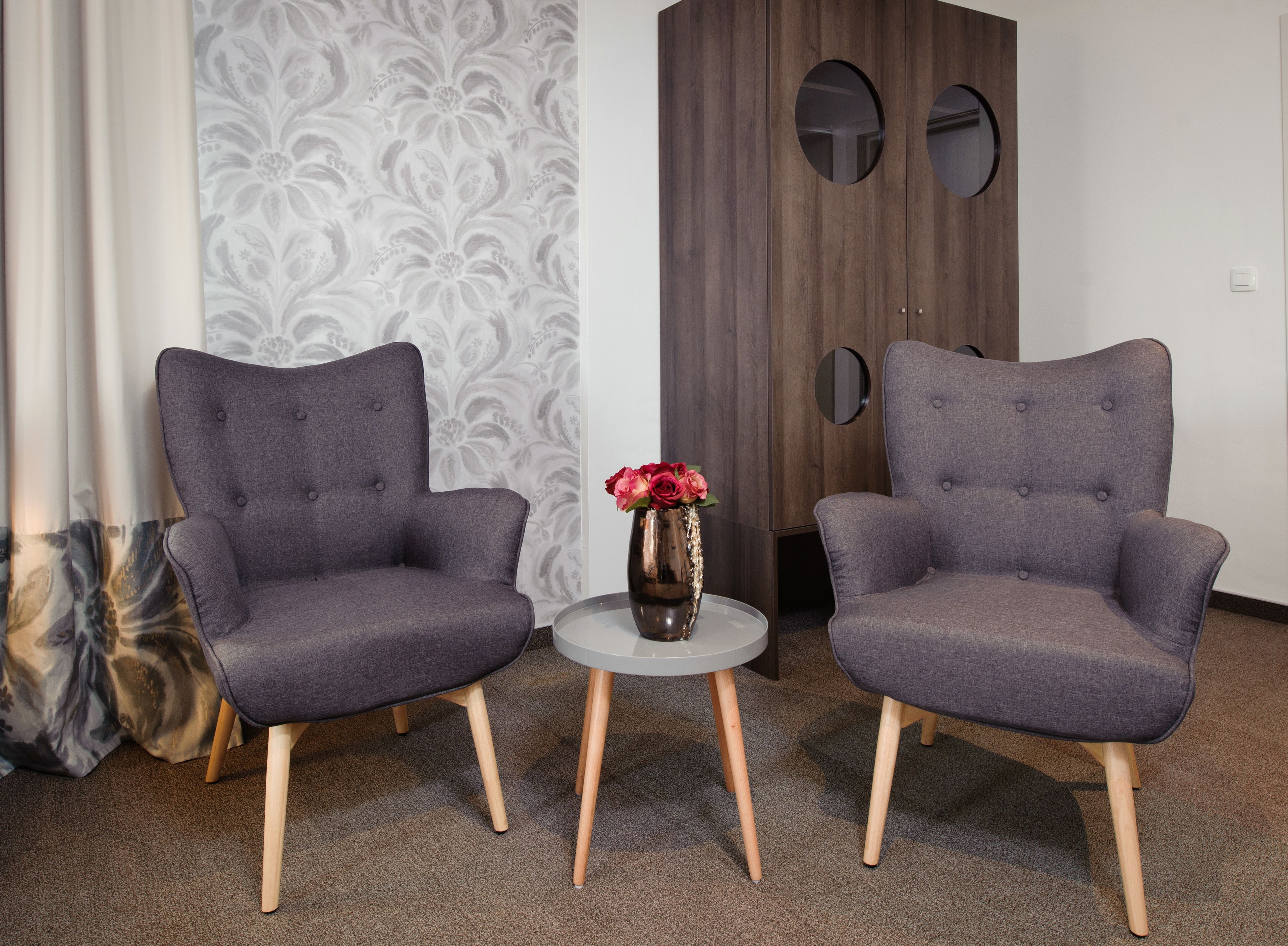 Dos sillones grises tapizados, patas de madera, una pequeña mesa redonda con un florero y una pared con papel pintado estampado.