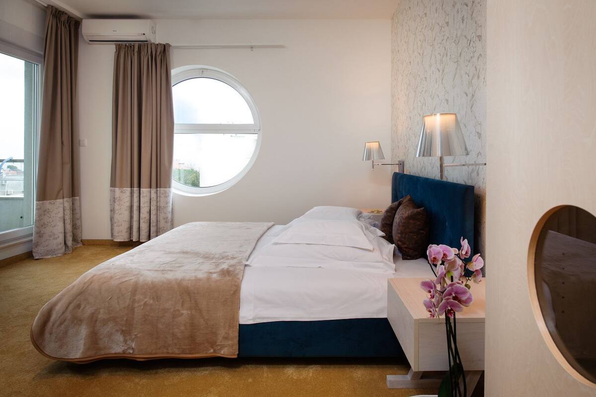 A hotel room with a bed, a round window, a nightstand with a lamp, and a wall-mounted mirror.